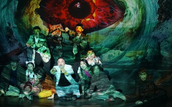 Stage scene from "Chuyến Đi Định Mệnh" with characters and a giant eye backdrop.